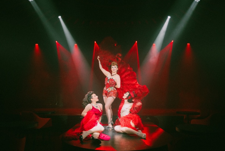 Three performers on stage wearing red burlesque costumes, stage lit with red lights.