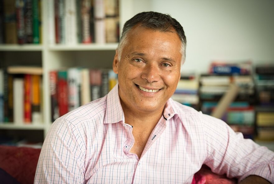 Journalist and author Stan Grant, a middle-aged man with short dark grey hair who is smiling, wearing a collared shirt and seated in front of a bookshelf.