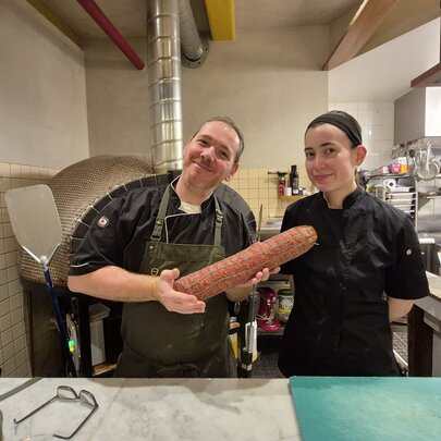 A chef and sous chef presenting a large hand-made salame in a chef's kitchen.