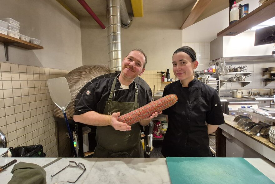 A chef and sous chef presenting a large hand-made salame in a chef's kitchen.