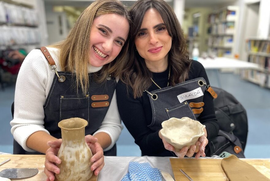 Two smiling Italian women in a pottery workshop  wearing aprons, proudly showing their clay pottery creations.