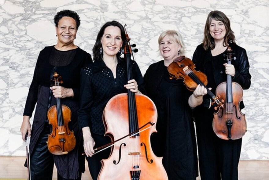 Four female classical musicians holding stringed instruments, wearing black, standing against a grey marble wall.