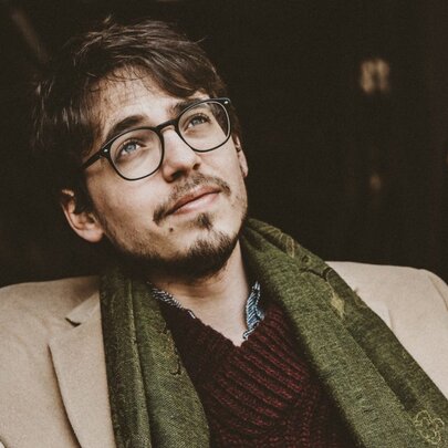French pianist, Lucas Debargue, reclining back in a chair, wearing glasses, a beige jacket, maroon knitted jumper and an olive green scarf.