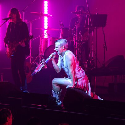 A performer lunging forward on a purple-lit stage under a white spotlight, a guitarist visible in the shadows.