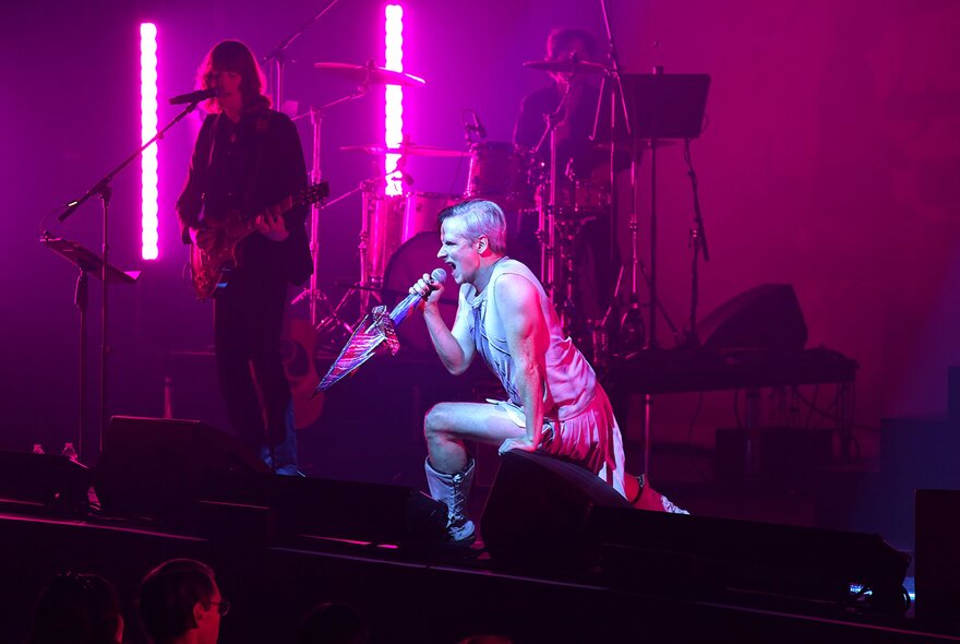 A performer lunging forward on a purple-lit stage under a white spotlight, a guitarist visible in the shadows.