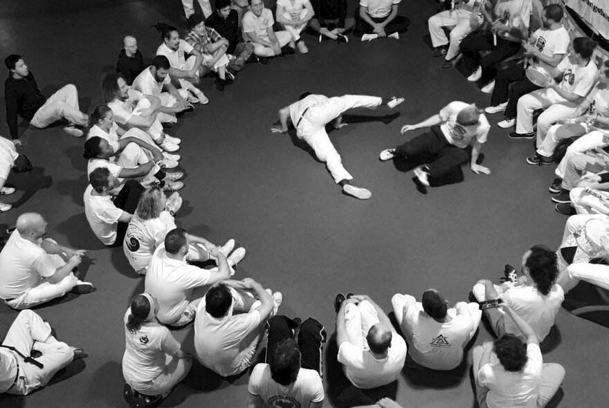 A black and white image of people demonstrating the Afro-Brazilian martial art of Capoeira in a small room with people sitting in a circle on the floor looking on. 