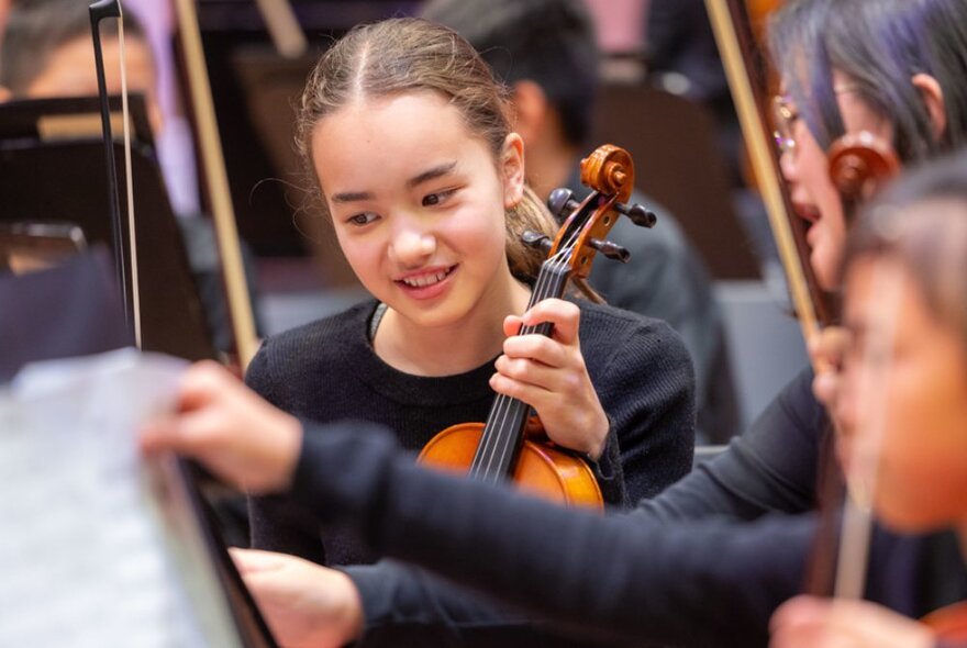 A young violinist, on stage with the Melbourne Youth Ochestra, two other young musicians blurred in the foreground.