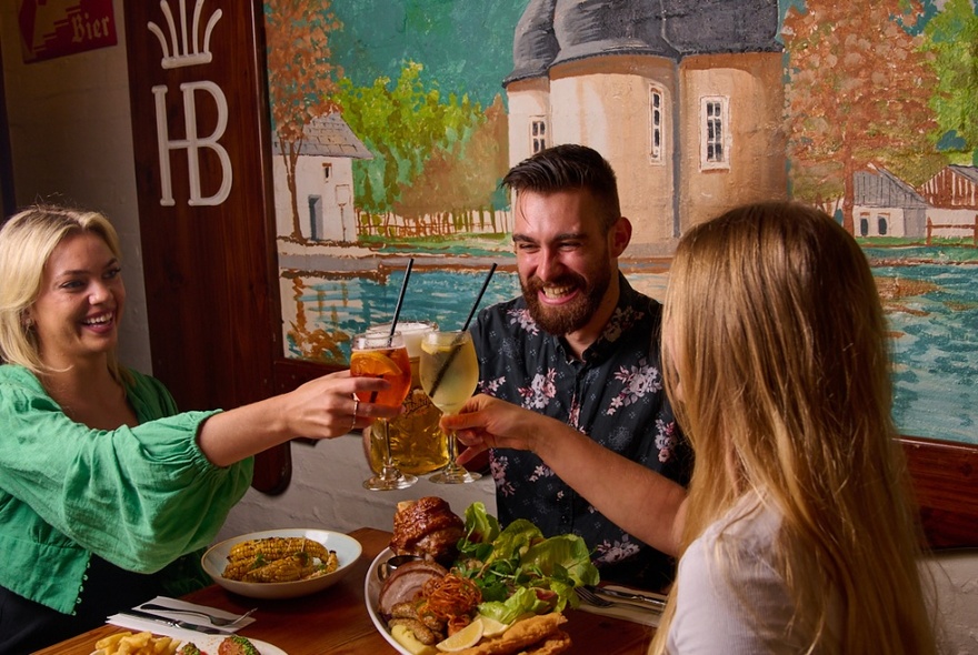 Three people raising their filled glasses for a toast, over a table of plates of food inside a restaurant with a mural on the wall behind them.