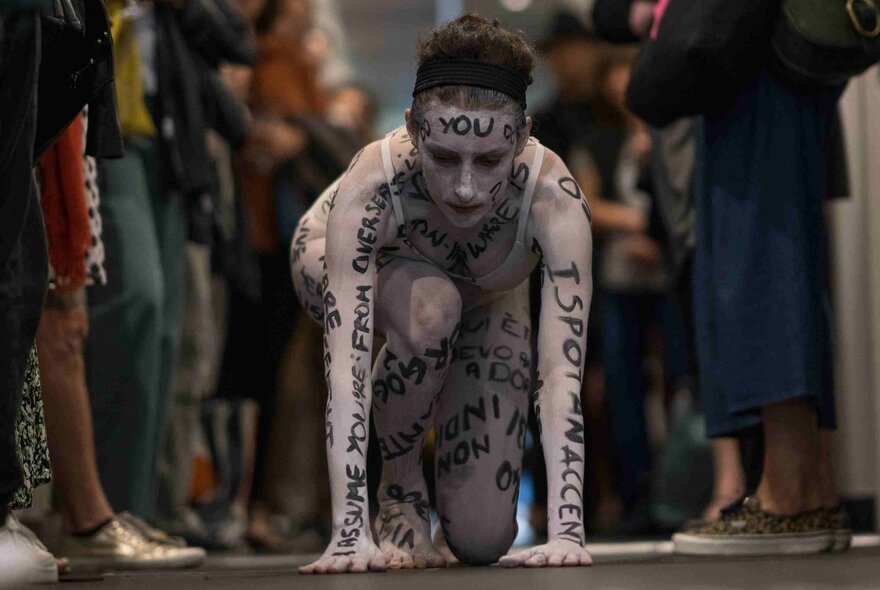 A woman who has painted her skin white and written black words all over her body, crouching on the ground in between two rows of people.
