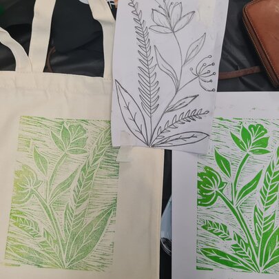 A green linocut of a flower and foliage printed on paper and a fabric tote bag.
