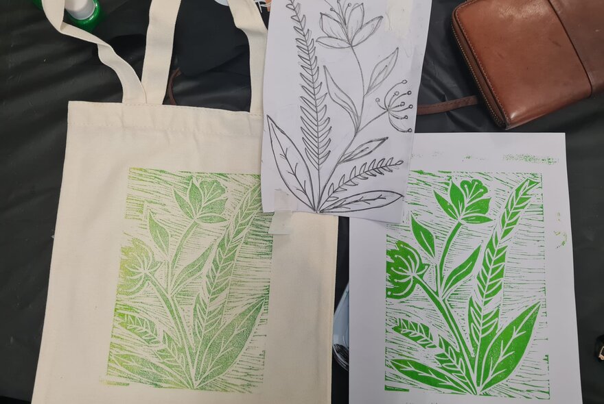 A green linocut of a flower and foliage printed on paper and a fabric tote bag.