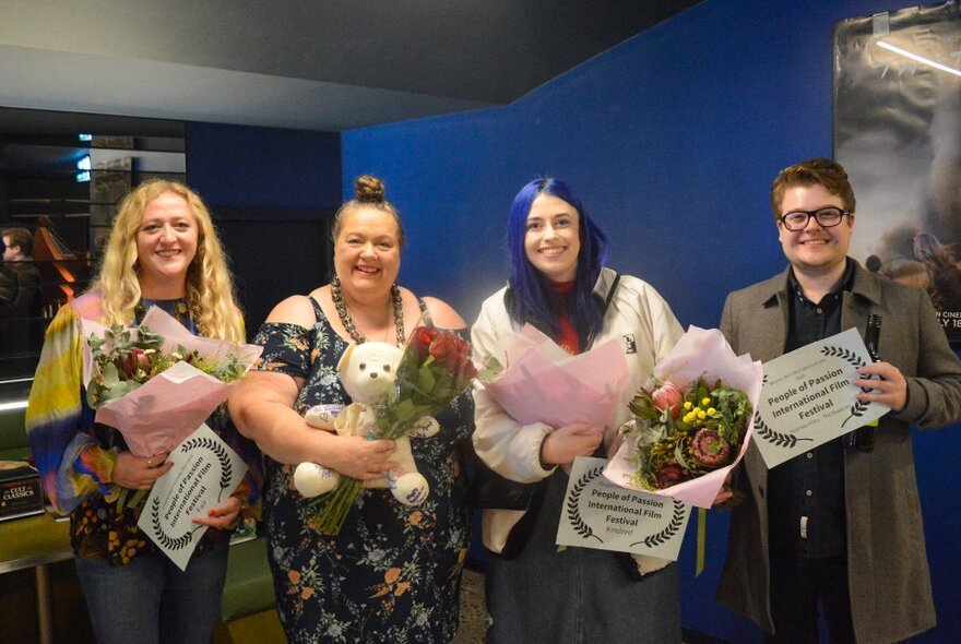 Four smiling people posed with bunches of flowers and holding certificates in their hands, in the foyer of a cinema.