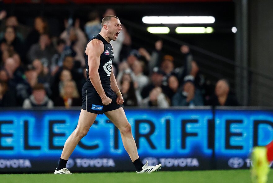 A Carlton AFL player celebrating on the field with his fists clenched by his sides, with blurred spectators behind him.