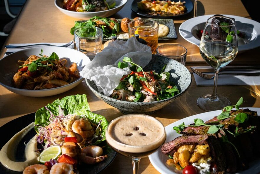 Several plates of food arranged on a table including a bowl of salad, meats, wedges and drinks.