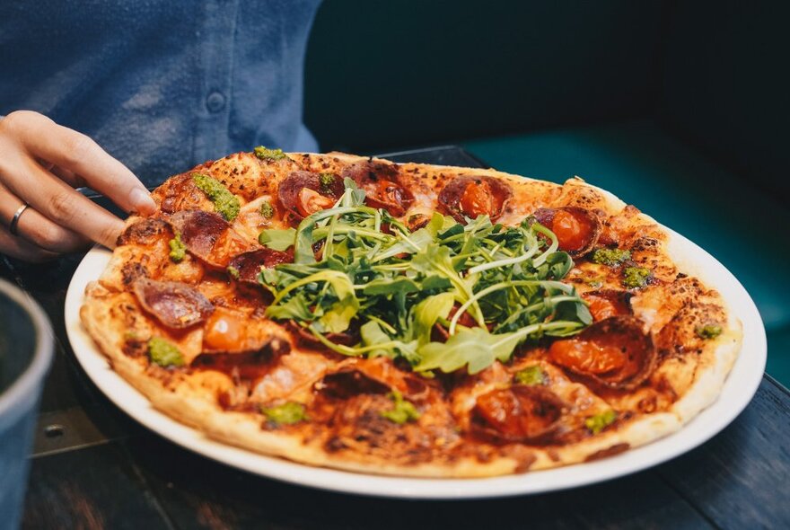 A hand on a plate containing a bubbly crusted pizza with rocket garnish. 