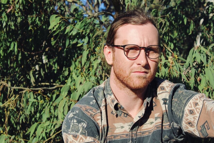 A portrait of Tim Loveday, a middle aged man outside sitting in front of a tree, wearing glasses and squinting slightly.