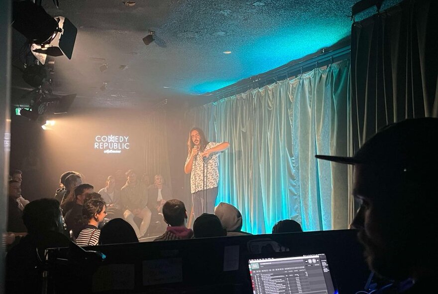 Stand-up comedian performing on a small stage in front of an audience.