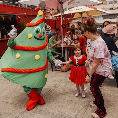 A person wearing a green, inflatable Christmas Tree mascot costume holding hands with a young child in a festive outdoor setting.