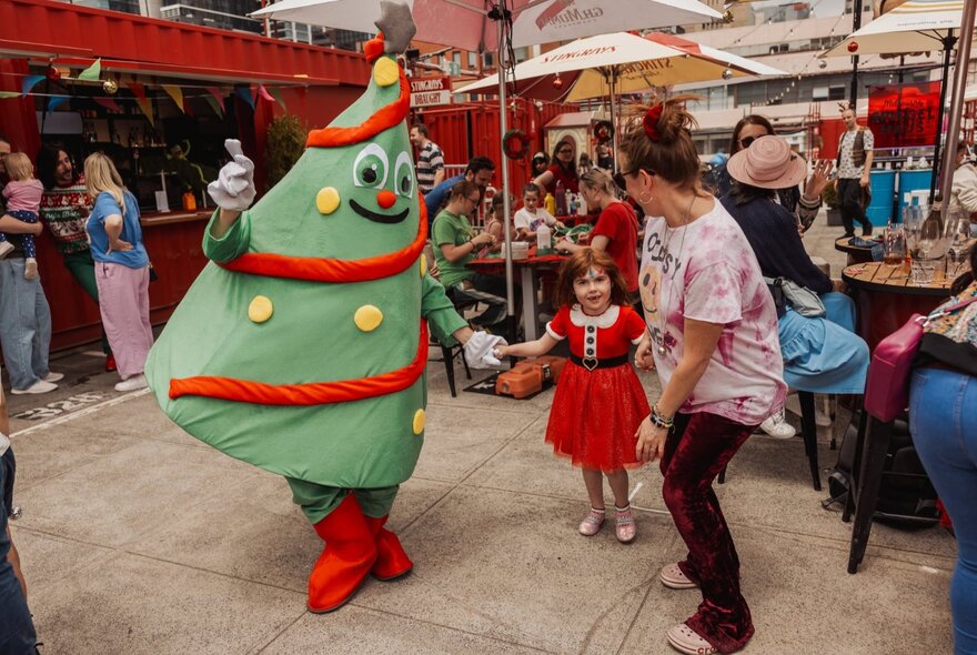 A person wearing a green, inflatable Christmas Tree mascot costume holding hands with a young child in a festive outdoor setting.