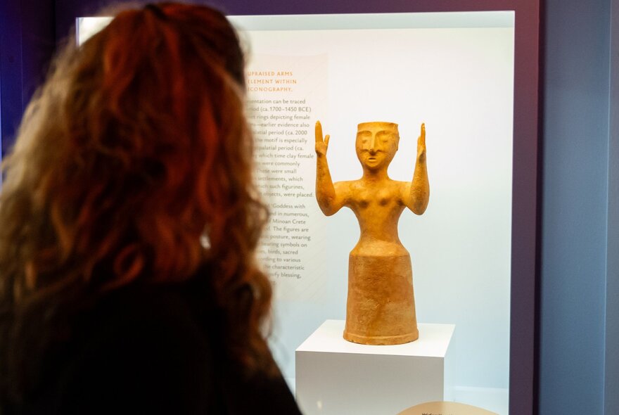 The back of a person's head looking at an small statue on display inside a glass case inside the Hellenic Museum.