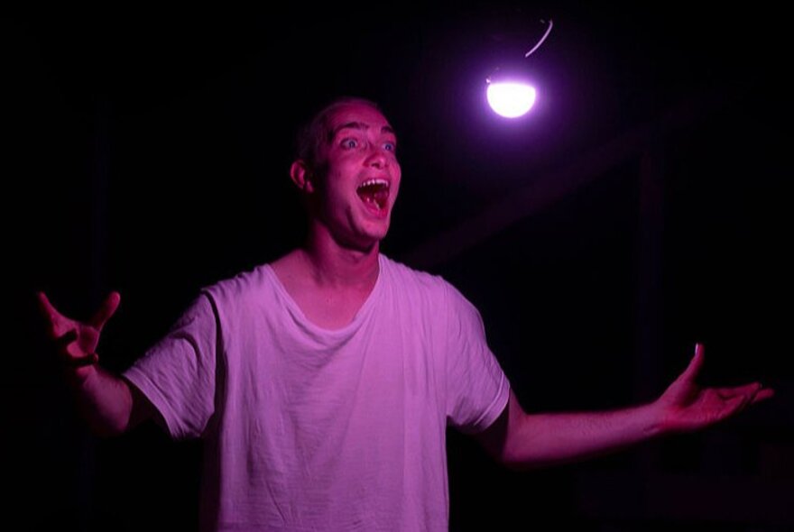 Physical theatre performer Daniel Nodder with his arms outstretched wearing a white t-shirt on a dark stage under a single white light globe.