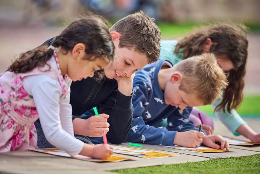 Four children sitting on the footpath drawing on paper, grass in the foreground.