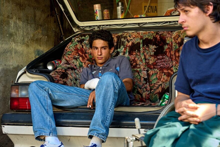 Still from a movie of a young man sitting in the open boot of a car, his legs dangling over the bumper bar, with another young person seated in the foreground.