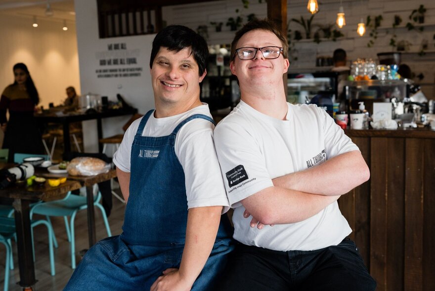 Two hospitality workers smiling and standing back to back inside a cafe.