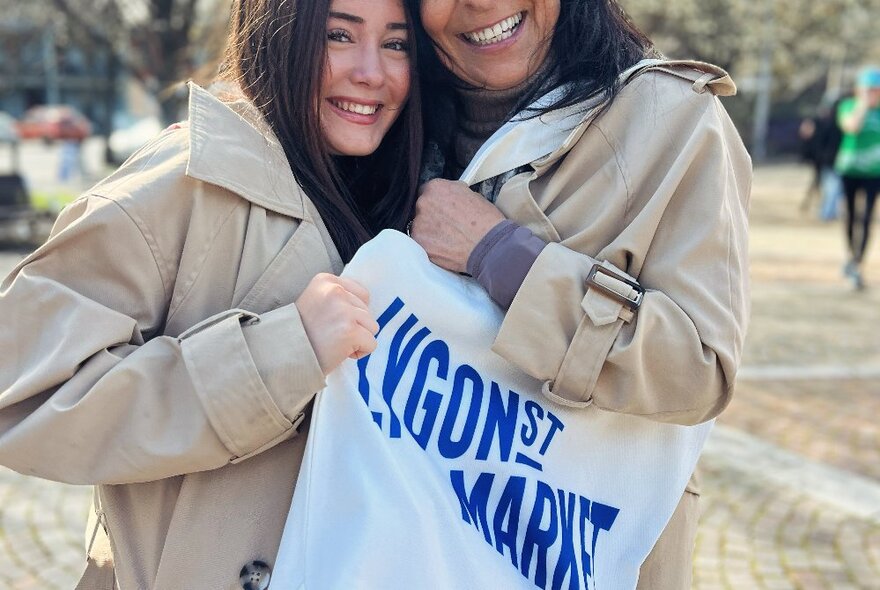 Two people holding up a calico tote bag with the words Lygon St Market printed on it.