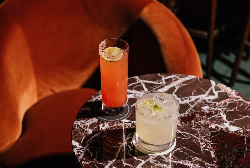 Two cocktails on a small round marble table, with a rust-coloured velvet chair in the background.