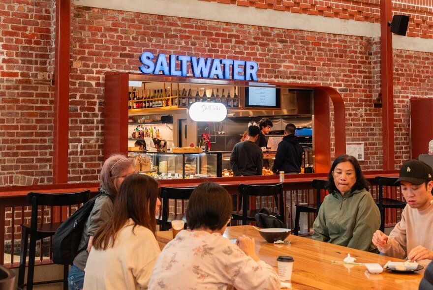 People eating at a wooden food court table in front of the Saltwater Sushi & Oyster Bar food stall at Queen Victoria Market.