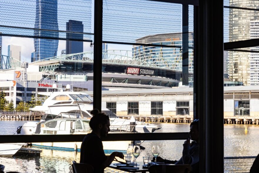 Dinser seated in silhouette at a table beside a panoramic window overlooking a harbour with boats and towers.