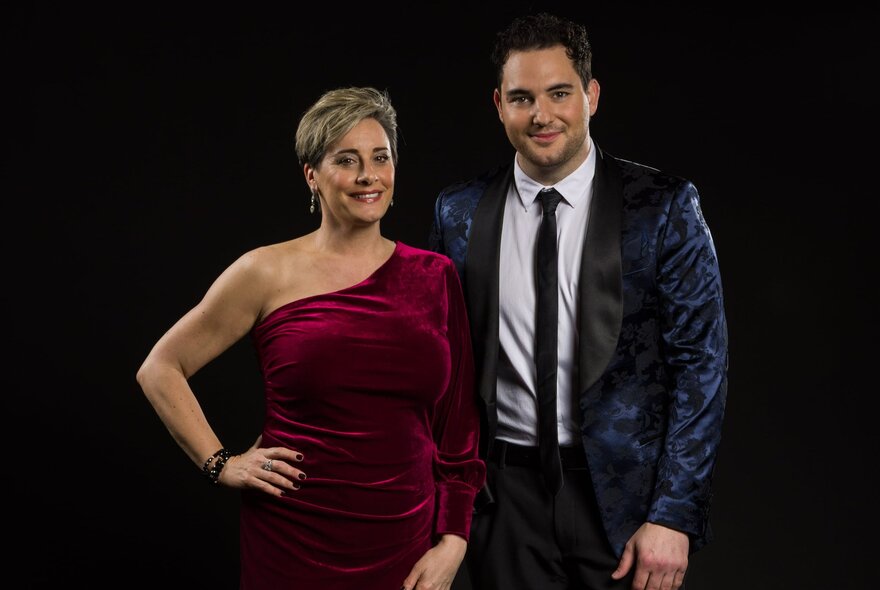 Singers Rhys Tolhurst and Nina Ferro wearing evening dress, posed in front of a black backdrop.