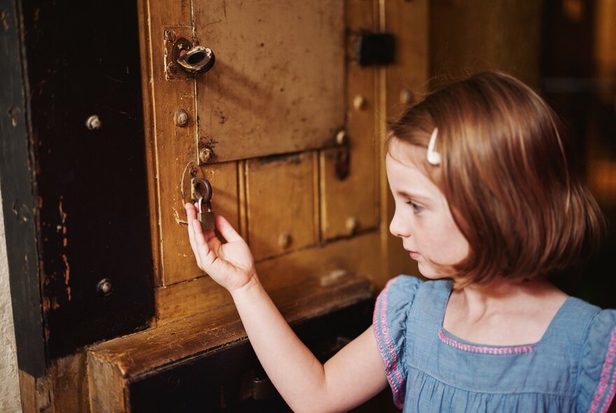 A young girl with bobbed hair picks at a padlock on an old prison cell door. 