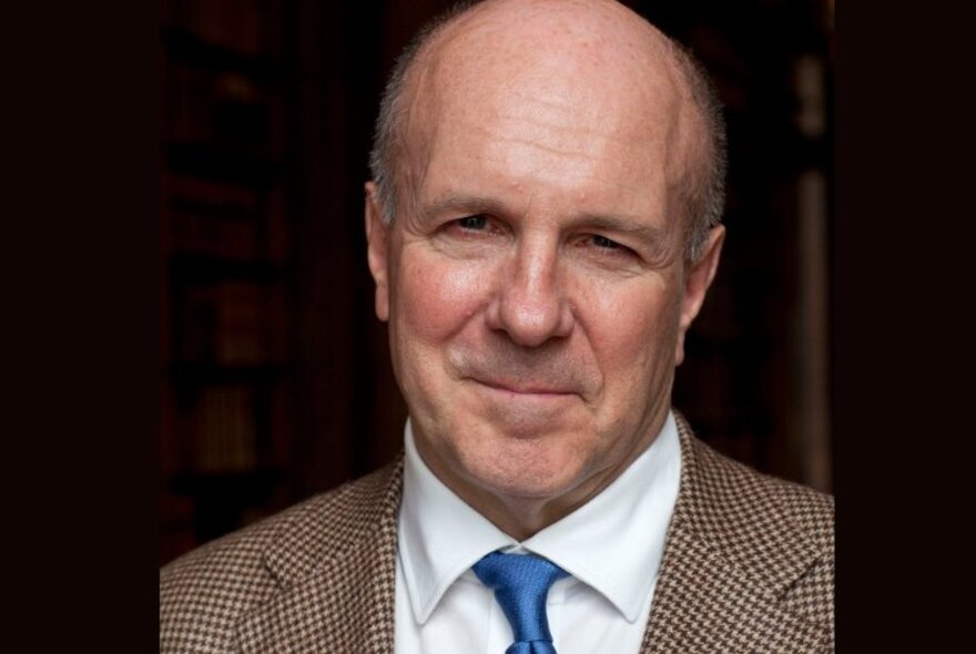 Portrait of author, Simon Winchester, subtly smiling at the camera and wearing a tweed jacket, white shirt and blue tie.