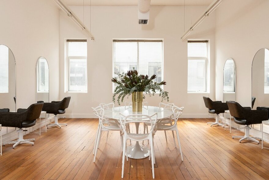 Interior of white room, high ceilings, black chairs facing mirrors on walls at side; large windows at rear; table with large bouquet of flowers at centre.