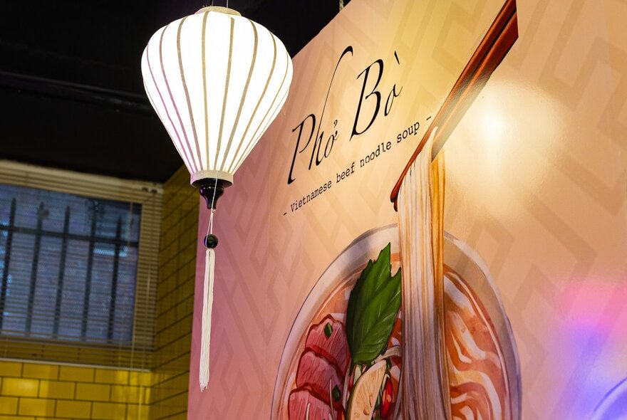 A white paper lantern in front of a wall-size mural of Pho Bo, Vietnamese beef noodle soup.