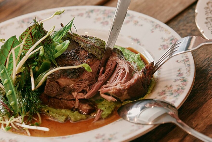 Knife and fork tucking into a slice of meat and vegetables on a floral decorated plate.