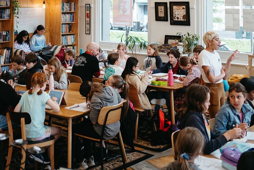 Children seated at worktables working with pens, paper and laptops.