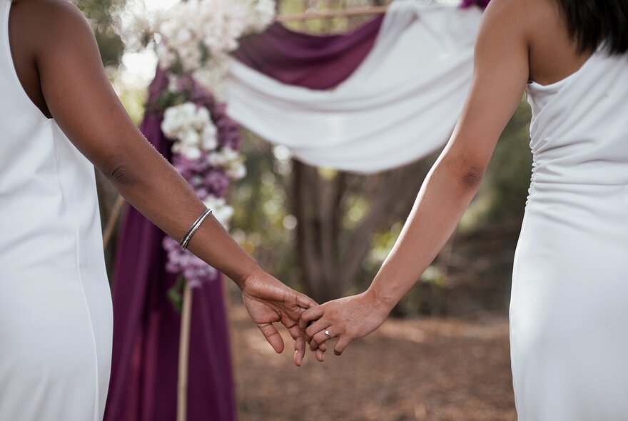 Two brides, each wearing white dresses, holding hands at an outdoor ceremony.