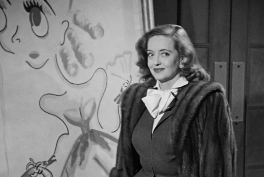 Black and white still from a film showing a woman with wavy hair wearing a fur jacket.