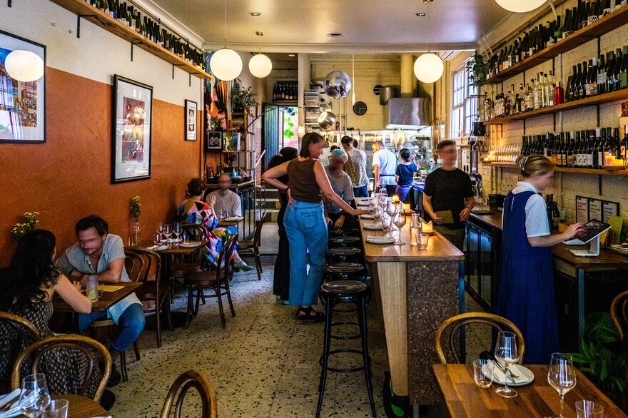 Inside a busy cosy wine bar people are seated drinking wine and the staff are behind the bar pouring wine and preparing food.