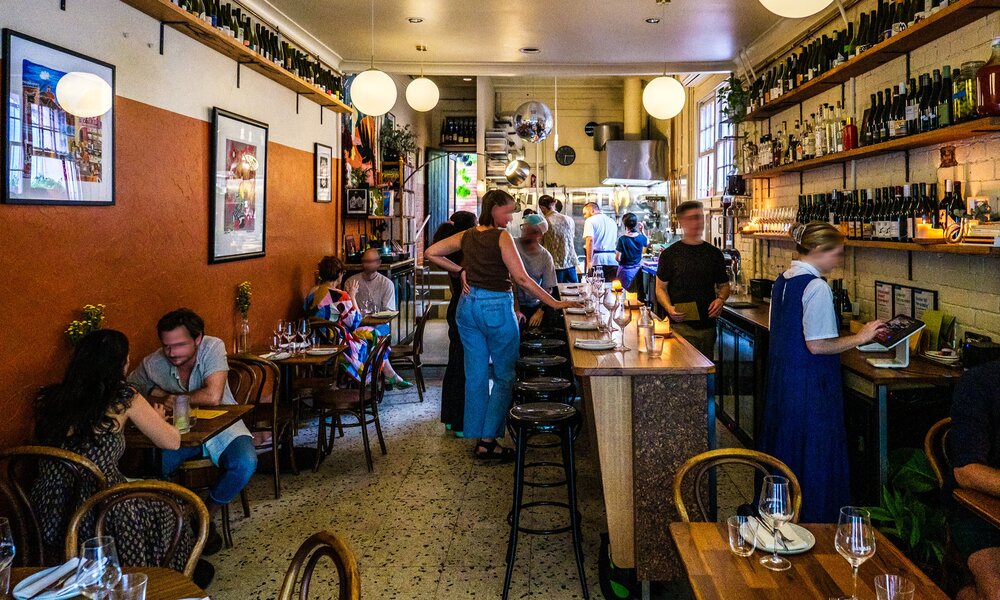 Inside a busy cosy wine bar people are seated drinking wine and the staff are behind the bar pouring wine and preparing food.