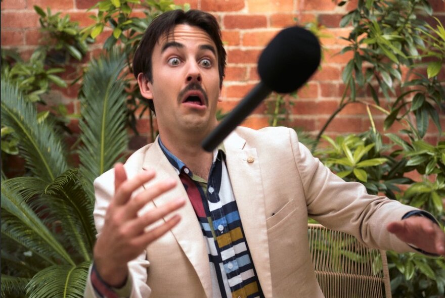 Comedian Sweeney Preston wearing a white suit jacket and throwing a reporter's microphone between his two hands. 