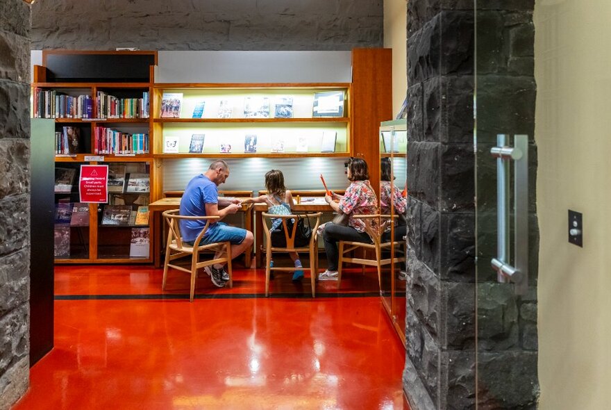 Looking through a bluestone walled hallway with a red-brown floor to three people at a table interacting. 