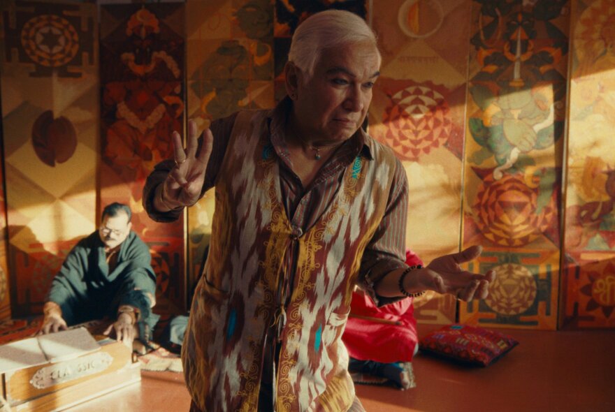 An older Indian man dancing in a sunlit room with colourful Indian motifs on the wall behind him.