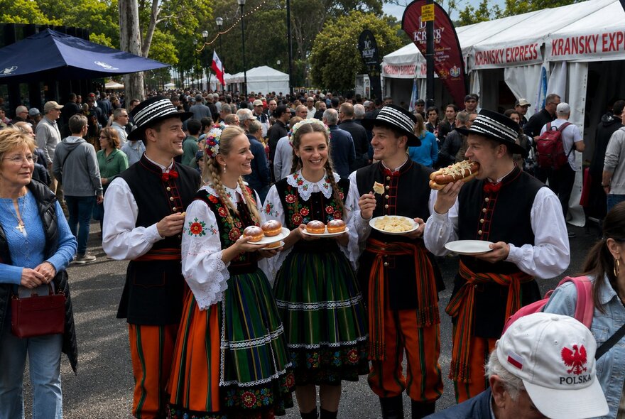People in Polish traditional dress at a festival, holding plates of food. 