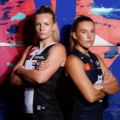 Two AFLW players standing back to back with arms crossed, one wearing a St Kilda jersey and the other in a Carlton jersey.