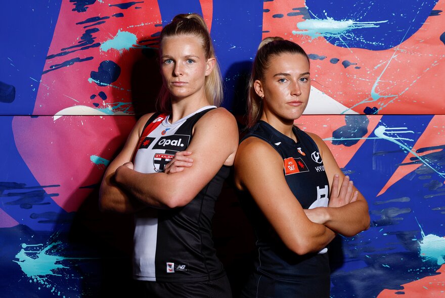 Two AFLW players standing back to back with arms crossed, one wearing a St Kilda jersey and the other in a Carlton jersey.