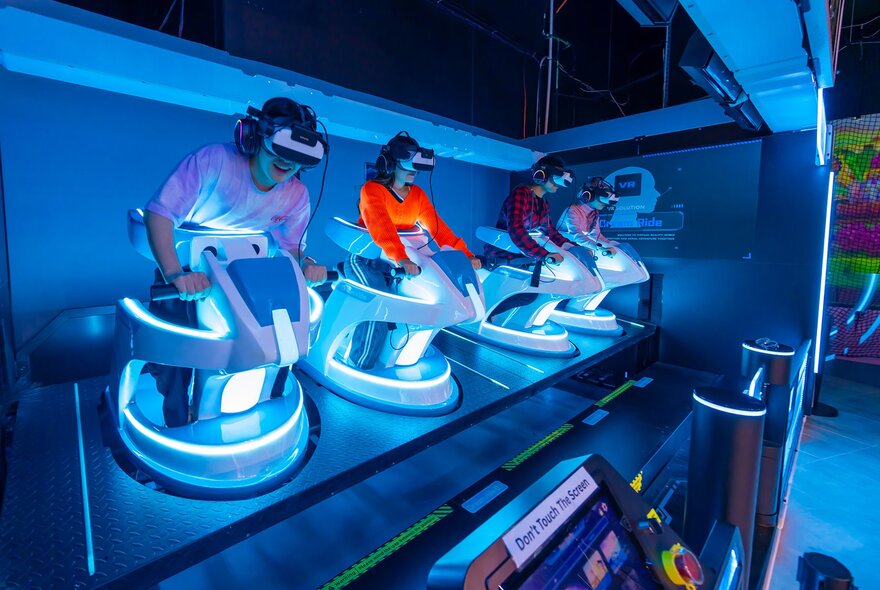 Four people riding pretend motorbikes with virtual reality headsets and blue lighting.